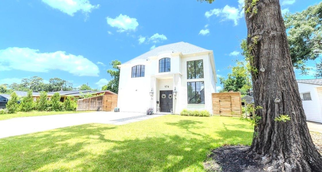 This photo showcases a modern two-story home with a clean white exterior and large windows, set in a lush green lawn. A spacious driveway leads to the entrance, and the property is framed by trees and a wooden fence, offering a bright and inviting curb appeal.
