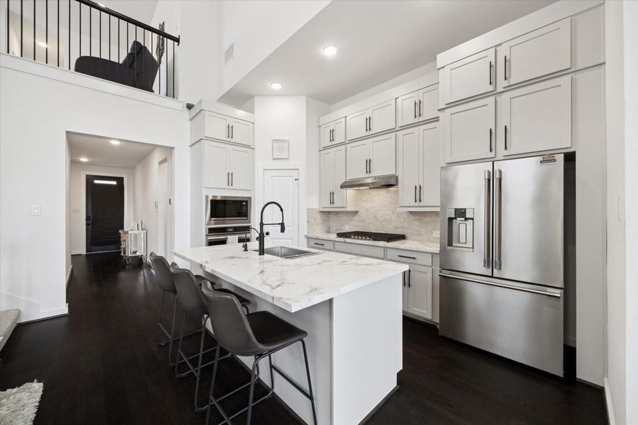 THE KITCHEN INCLUDES WHITE CABINETRY, A LARGE CENTER ISLAND WITH A SINK, AND STAINLESS STEEL APPLIANCES INCLUDING A FRENCH-DOOR REFRIGERATOR.