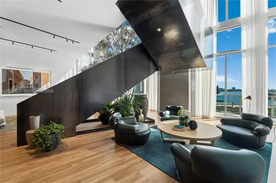 Sculptural black metal staircase, double-height floor-to-ceiling windows, wood-finish flooring, and recessed ceiling track lighting