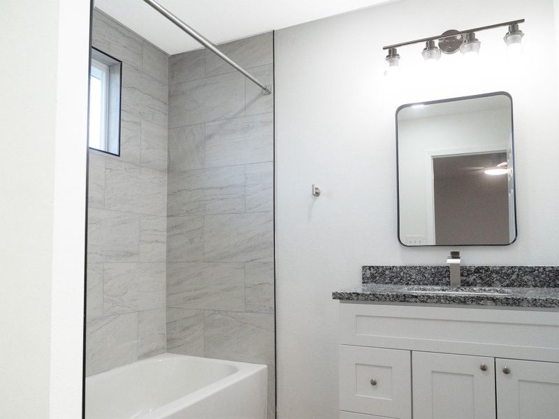 Modern bathroom featuring a sleek vanity with granite countertop, framed mirror, and stylish light fixture. The shower area has elegant gray tile and a window for natural light.
