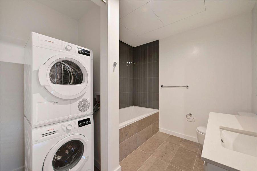 Sleek and functional bathroom featuring a modern soaking tub with vertical tile surround, neutral stone flooring, and a clean white vanity. A stacked Bosch washer and dryer set is conveniently located just off the bathroom, offering an efficient and space-saving laundry solution.