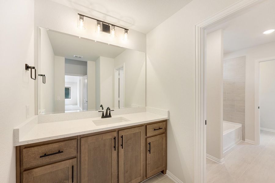 Bathroom vanity with wood cabinets, white countertop, large mirror, and tiled shower area visible through doorway Bathroom vanity with wood cabinets, white countertop, large mirror, and tiled shower area visible through doorway