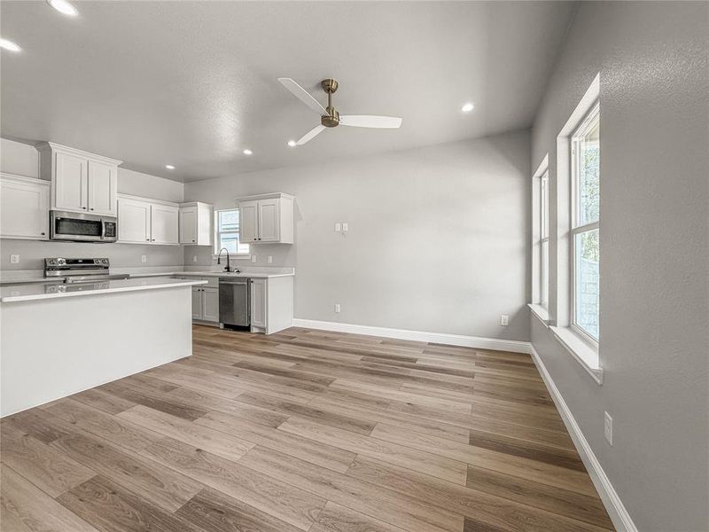 Open-concept living space featuring wood-finish flooring, a white kitchen island, stainless steel appliances, and multiple windows Open-concept living space featuring wood-finish flooring, a white kitchen island, stainless steel appliances, and multiple windows