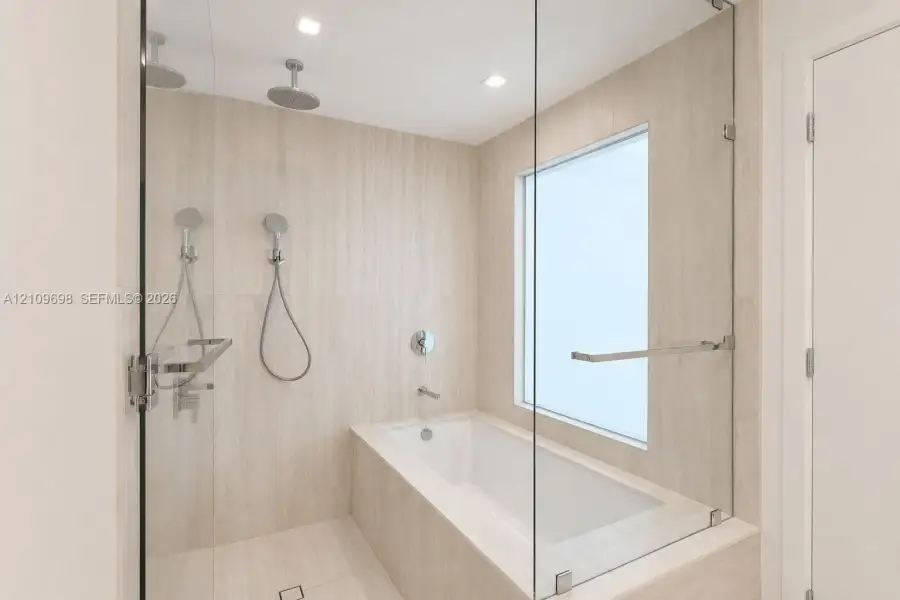 Enclosed wet room featuring a deep soaking tub, frameless glass partition, dual wall-mounted rainfall showerheads, and neutral-toned stone-finish tiling