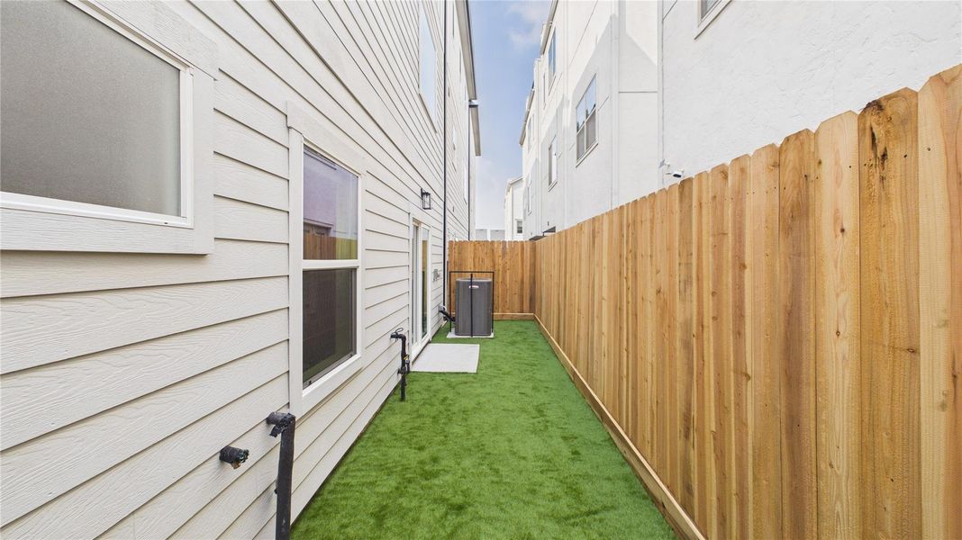 This photo shows a narrow side yard with artificial grass between two buildings. It features a wooden fence on one side and the exterior wall of a house with windows on the other. An air conditioning unit is visible at the end of the yard.