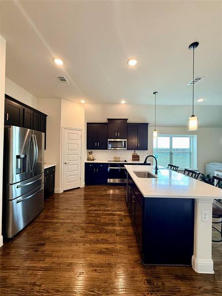 Kitchen featuring dark wood-finish cabinetry, stainless steel appliances, a spacious island with a white countertop and integrated sink, and recessed lighting Kitchen featuring dark wood-finish cabinetry, stainless steel appliances, a spacious island with a white countertop and integrated sink, and recessed lighting