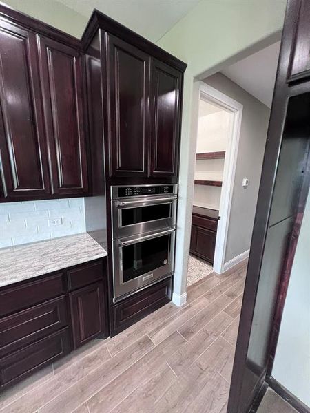 Kitchen with stainless steel double oven, tasteful backsplash, light wood-type flooring, and dark brown cabinets