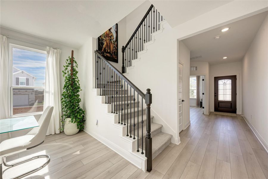 Entry foyer featuring light wood-finish flooring, a dark wood entry door with decorative glass, recessed lighting, and a staircase with carpeted treads and black metal balusters