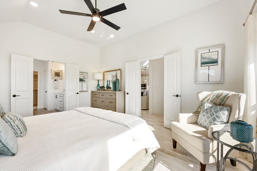 Bright master bedroom with ceiling fan, light wood floors, sitting chair, and doors leading to walk-in closet and bathroom