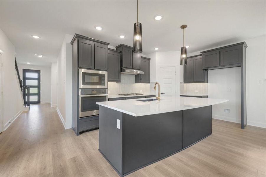 Modern kitchen featuring dark cabinetry, stainless steel appliances, a white island with pendant lighting, and wood-finish flooring