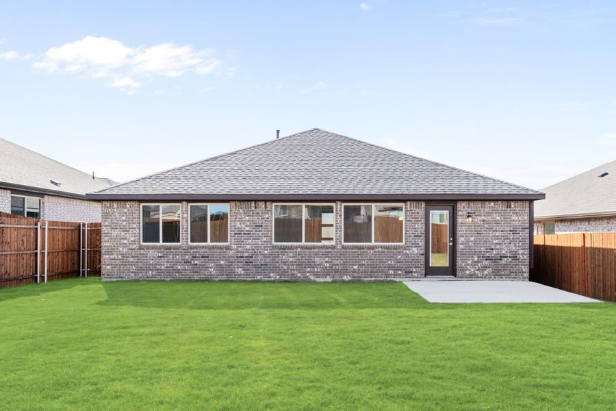 Rear view of single-story brick home with large windows, concrete patio, and fenced green backyard Rear view of single-story brick home with large windows, concrete patio, and fenced green backyard