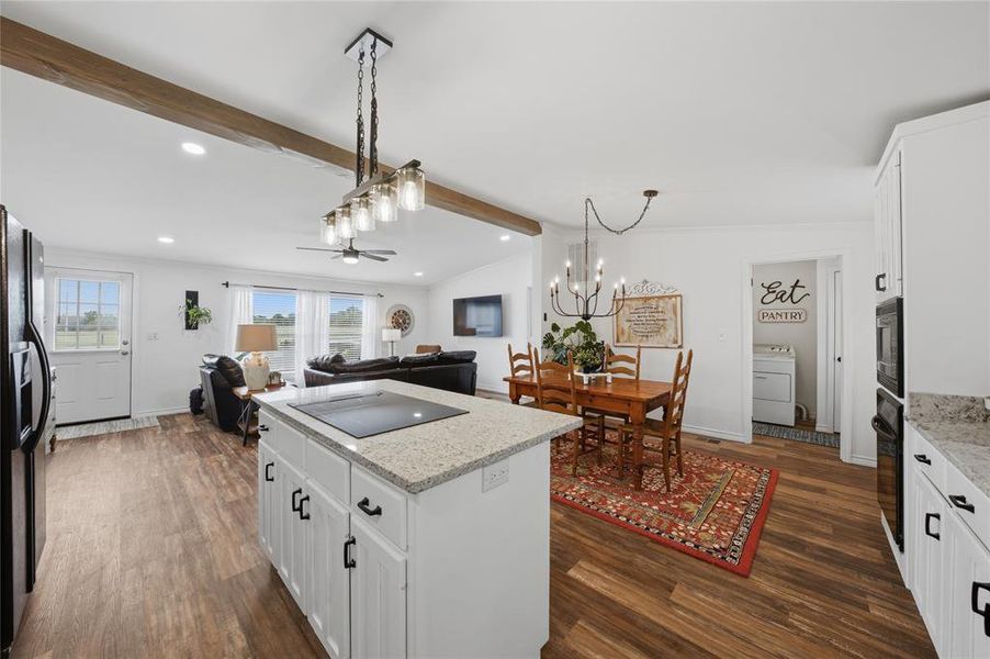 Open-concept living area featuring wood-finish flooring, exposed ceiling beams, white cabinetry with black hardware, granite countertops, and a built-in oven