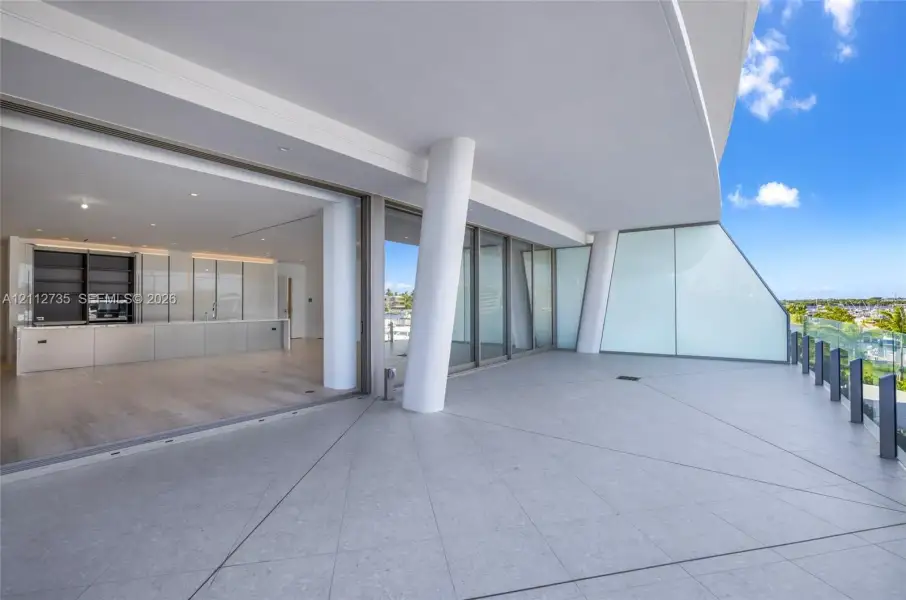 Expansive covered terrace featuring large-format tile flooring, structural pillars, and glass railings, with interior space showcasing wood-finish flooring and built-in cabinetry