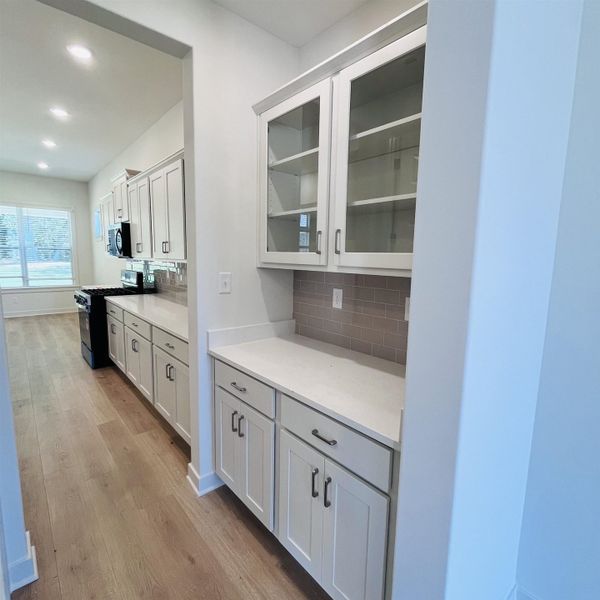 White shaker cabinetry, light wood-finish flooring, recessed lighting, subway tile backsplash, and built-in glass-front upper cabinets, home also features a full walk in pantry as well