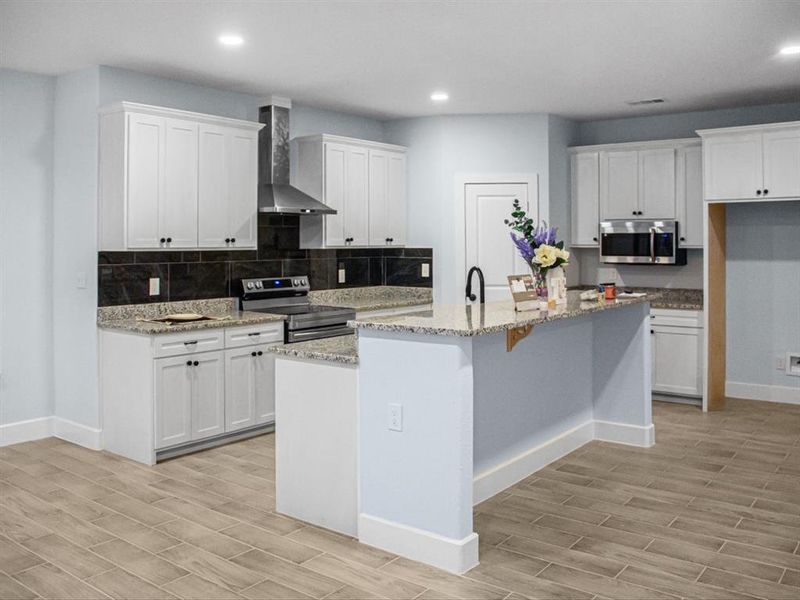 Kitchen with stainless steel appliances, wall chimney range hood, a kitchen island with sink, white cabinetry, and recessed lighting