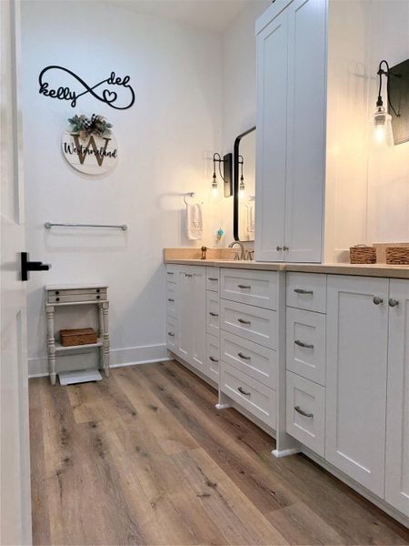 This bathroom features a modern design with ample storage in white cabinets, a wooden countertop, and stylish lighting fixtures. The light wood flooring and decorative wall accents add warmth and charm.