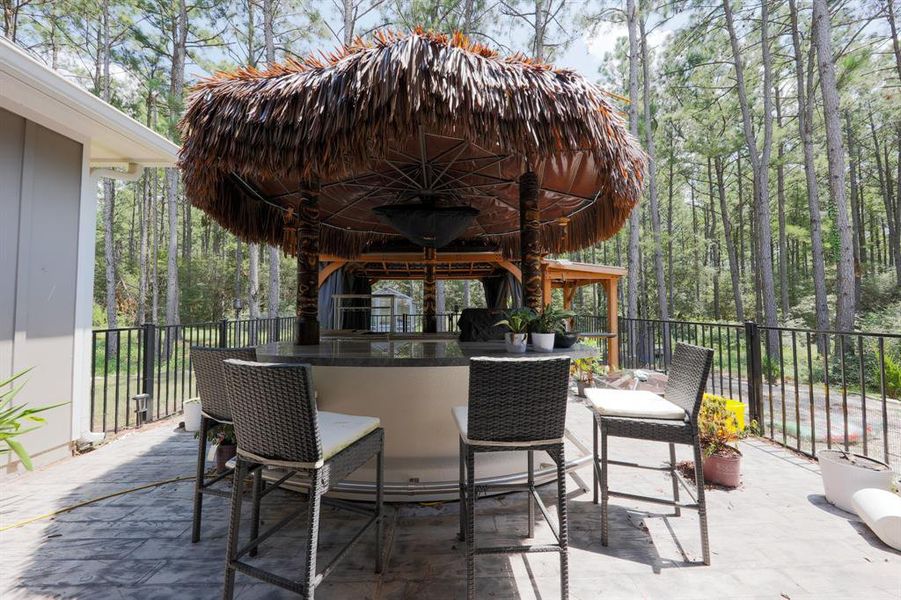 This photo showcases a spacious outdoor patio area with a tiki-style bar, surrounded by lush greenery and tall trees. The space features bar seating, a thatched roof, and a fenced perimeter, perfect for entertaining and enjoying nature.