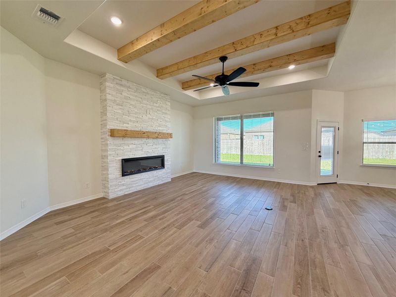 Unfurnished living room featuring a tray ceiling, light wood-style flooring, beamed ceiling, a stone fireplace, and a ceiling fan