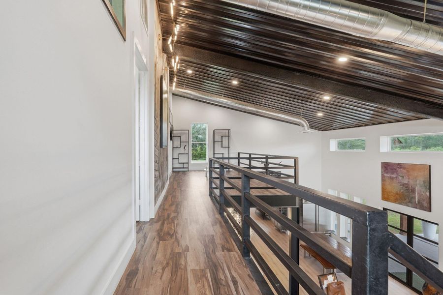 This photo showcases a modern, industrial-style interior with a high ceiling and exposed metal ductwork. The space features a mezzanine walkway with sleek railings, wood flooring, and ample natural light from large windows, creating an open and airy atmosphere.