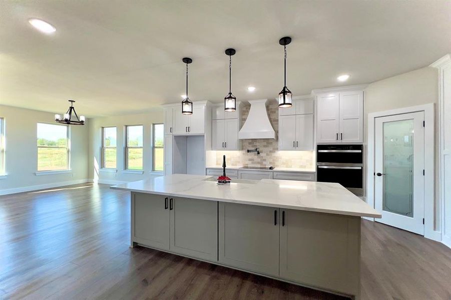 Kitchen featuring backsplash, hanging light fixtures, white cabinetry, custom range hood, and a kitchen island with sink
