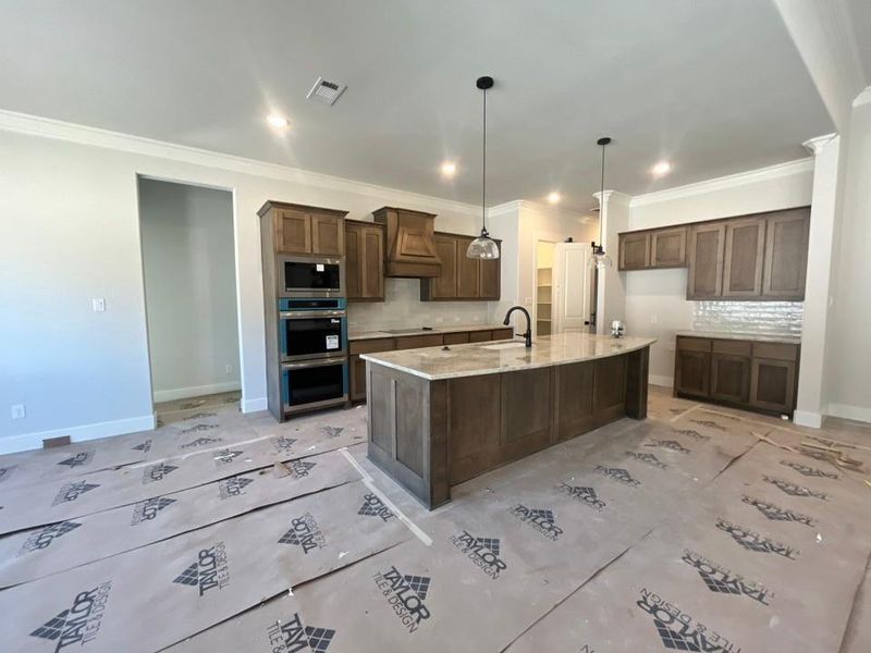 Kitchen featuring a center island with sink, light stone counters, tasteful backsplash, stainless steel appliances, and crown molding