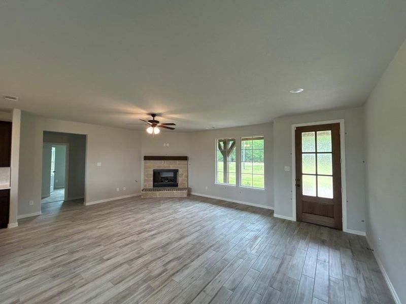Spacious open concept living area featuring wood-finish flooring, a corner fireplace with stone surround, large windows, and a frosted glass-paneled door Spacious open concept living area featuring wood-finish flooring, a corner fireplace with stone surround, large windows, and a frosted glass-paneled door