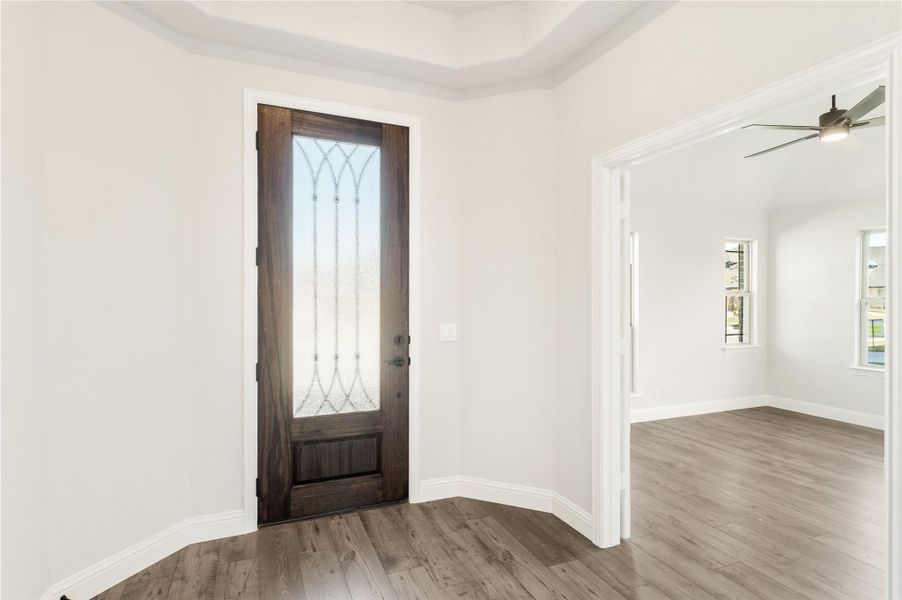 Entryway with dark wood front door featuring decorative glass panel and hardwood floors, opening to adjacent room with ceiling fan