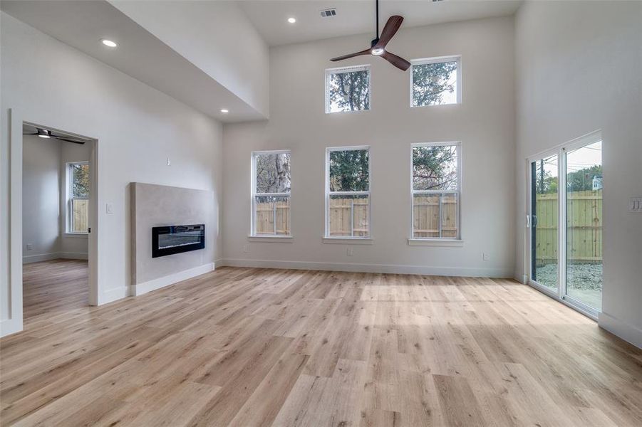 Unfurnished living room featuring ceiling fan, a high ceiling, light wood-type flooring, a glass covered fireplace, and healthy amount of natural light