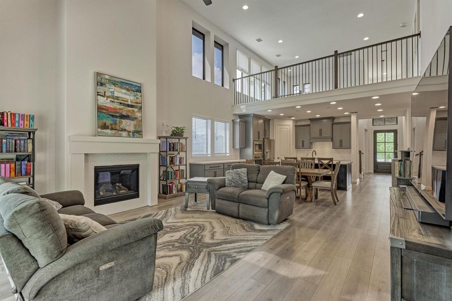 This spacious open-concept living area features high ceilings and abundant natural light. It includes a cozy fireplace, modern kitchen, and a stylish dining area, all with contemporary finishes. The loft above adds extra space and overlooks the living area, enhancing the home's airy feel.