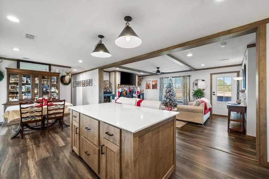 Open concept living area featuring wood-finish flooring throughout, a central kitchen island with a light-toned countertop, recessed lighting, and exposed wood beam accents