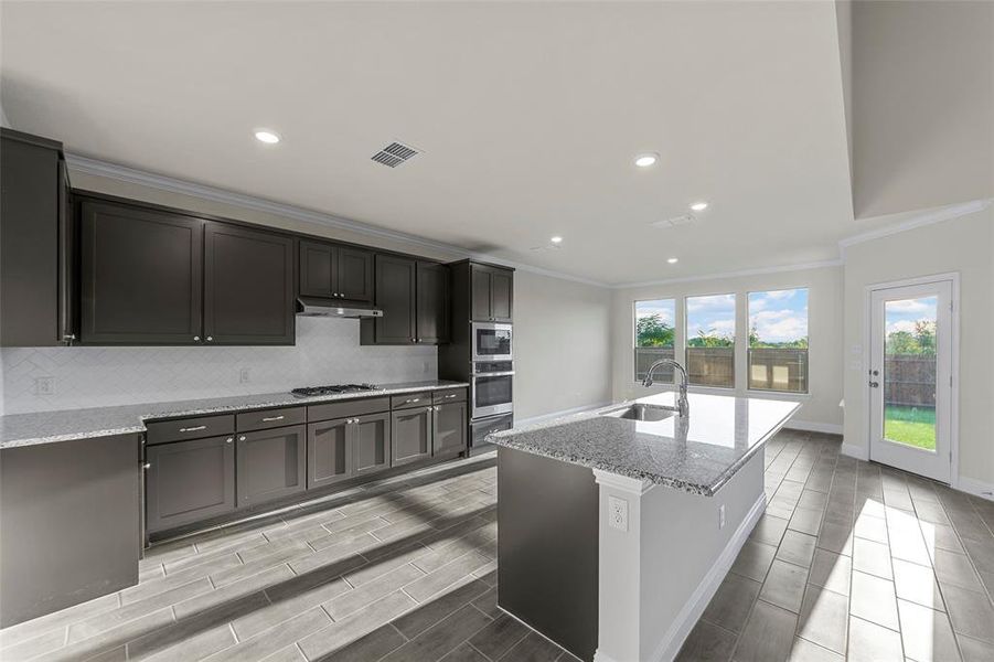 Kitchen with light stone counters, backsplash, ornamental molding, an island with sink, and recessed lighting Kitchen with light stone counters, backsplash, ornamental molding, an island with sink, and recessed lighting