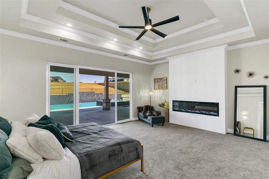 Carpeted bedroom featuring a large fireplace, ceiling fan, access to outside, a tray ceiling, and crown molding