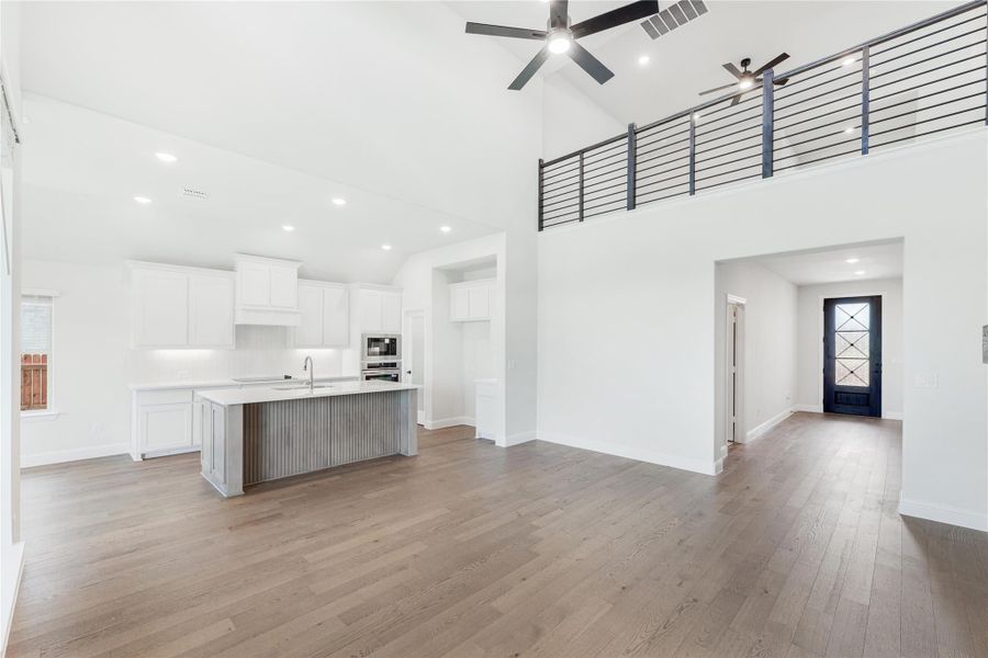 Open living area with high ceilings, hardwood floors, and white kitchen with center island and second-floor loft railing visible above.