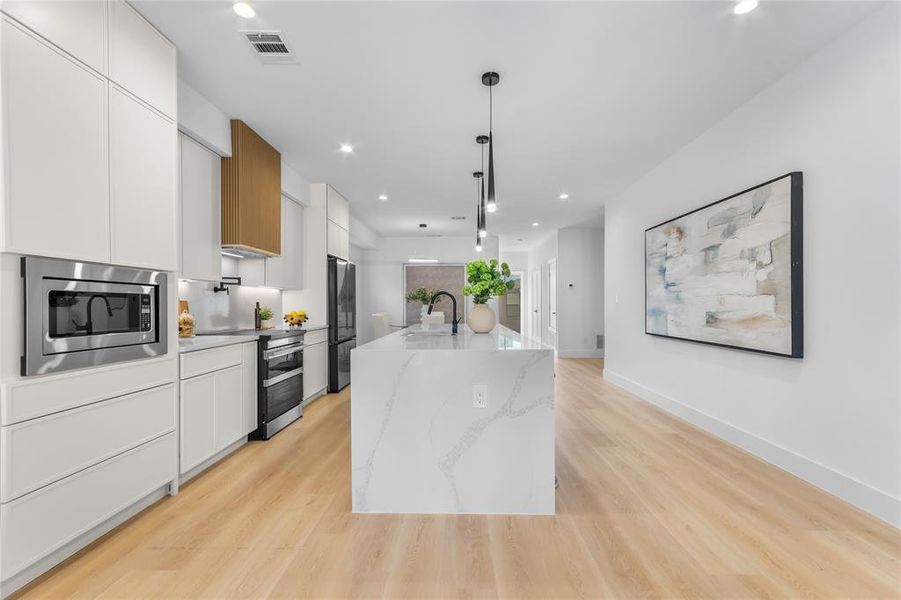 Contemporary kitchen featuring a spacious island with a waterfall-edge countertop, light wood-finish flooring, white flat-panel cabinetry, a stainless steel built-in microwave, and pendant lighting Contemporary kitchen featuring a spacious island with a waterfall-edge countertop, light wood-finish flooring, white flat-panel cabinetry, a stainless steel built-in microwave, and pendant lighting