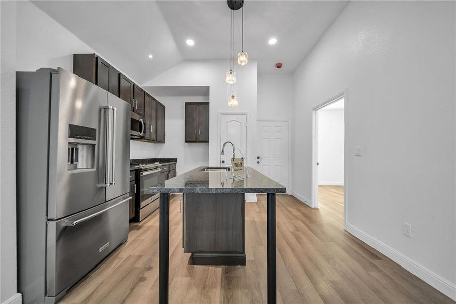 Kitchen featuring stainless steel appliances, dark brown cabinetry, decorative light fixtures, a center island with sink, and light wood-style floors
