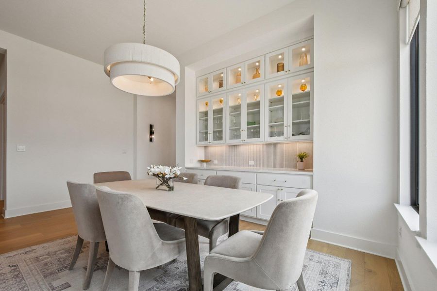 Dining area featuring light wood flooring, a decorative overhead light fixture, and a built-in cabinet unit with glass doors and accent lighting Dining area featuring light wood flooring, a decorative overhead light fixture, and a built-in cabinet unit with glass doors and accent lighting