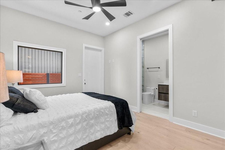 This is a modern bedroom with light walls and wood flooring, featuring a cozy bed, a ceiling fan, and a window. It includes an en-suite bathroom with a sleek vanity and toilet, providing convenience and style. This is a modern bedroom with light walls and wood flooring, featuring a cozy bed, a ceiling fan, and a window. It includes an en-suite bathroom with a sleek vanity and toilet, providing convenience and style.