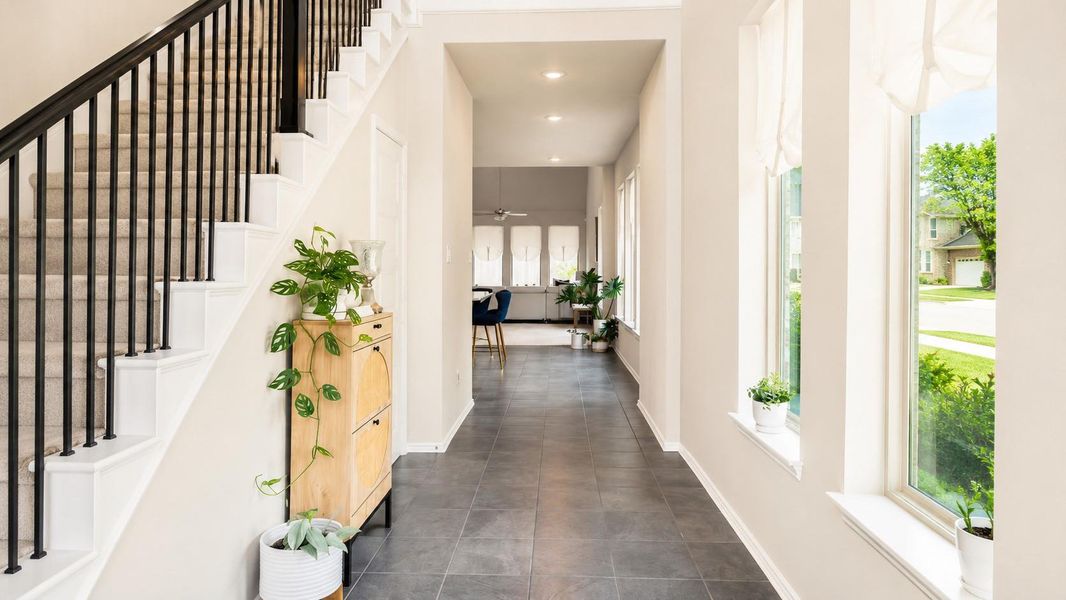 A wide hallway with dark gray tile flooring provides a direct view from the entryway into the rear living spaces. The area includes a carpeted staircase on one side and a series of large windows on the other.