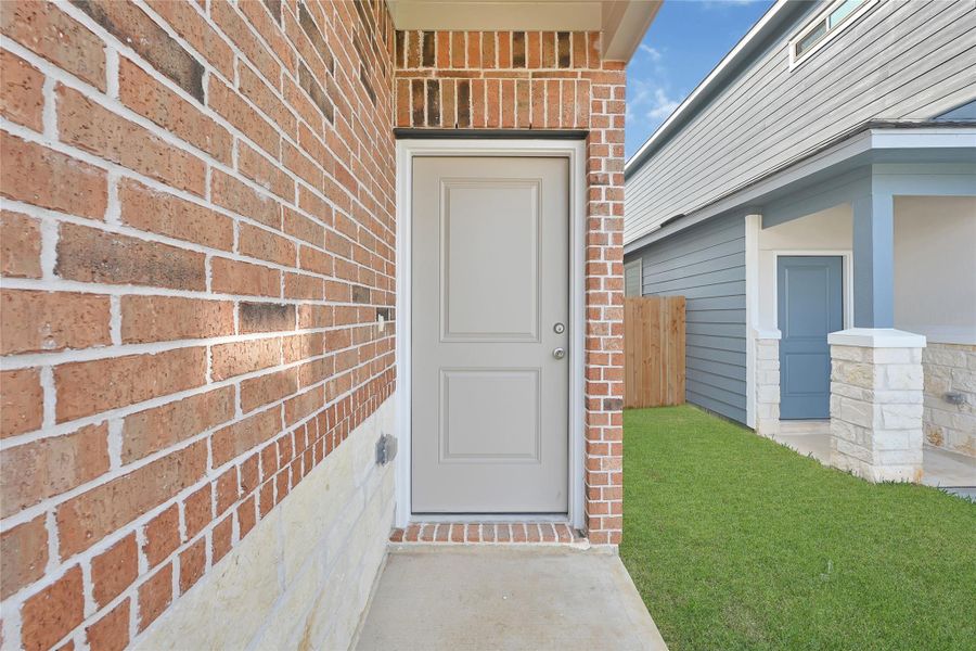 This photo shows a modern home entrance with a two-panel front door, surrounded by brick and stone. Covered Patio greens you in! Please note: Image is not the actual home; it is a home with the same floor plan in the neighborhood.