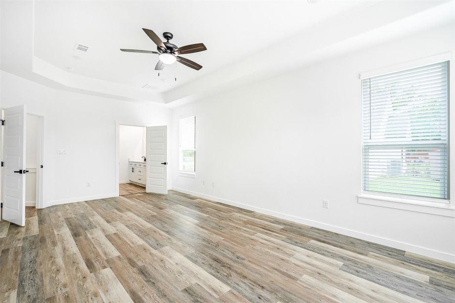 A spacious primary bedroom features tray ceilings, wood-look flooring, and ample natural light. A spacious primary bedroom features tray ceilings, wood-look flooring, and ample natural light.