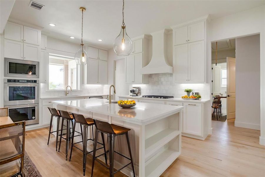 Kitchen featuring custom exhaust hood, stainless steel appliances, tasteful backsplash, a center island with sink, and light countertops Kitchen featuring custom exhaust hood, stainless steel appliances, tasteful backsplash, a center island with sink, and light countertops