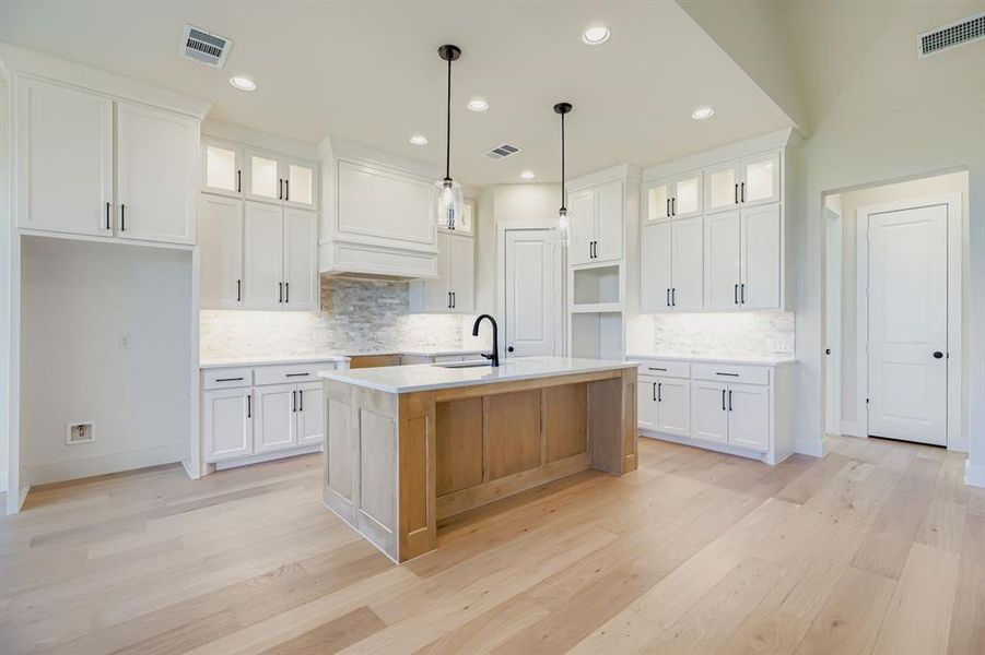 Kitchen with glass insert cabinets, white cabinetry, a kitchen island with sink, hanging light fixtures, and light wood-type flooring Kitchen with glass insert cabinets, white cabinetry, a kitchen island with sink, hanging light fixtures, and light wood-type flooring