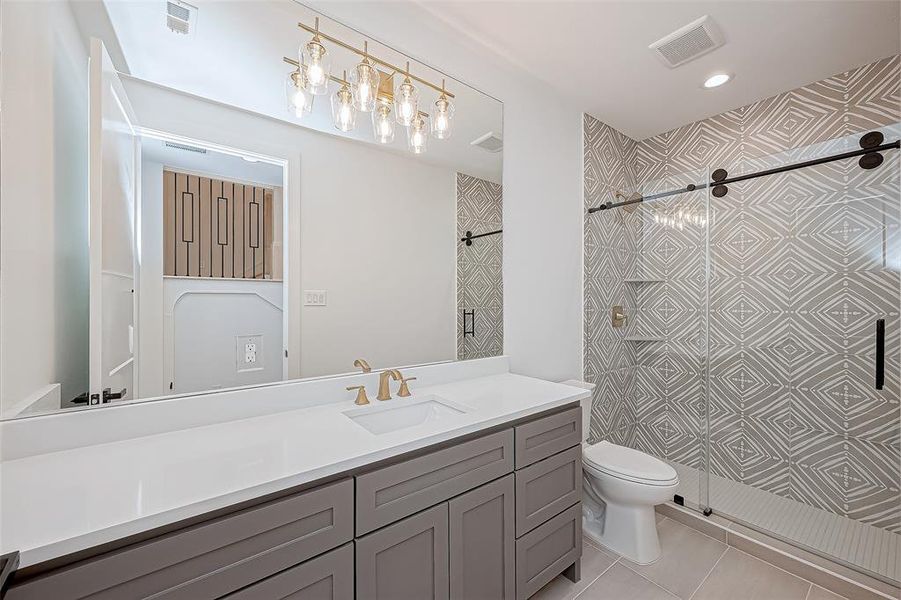 This bathroom features vanity with lots of storage,  gold fixtures, a large mirror, and stylish lighting. The walk-in shower has a geometric tile design and a sliding glass door