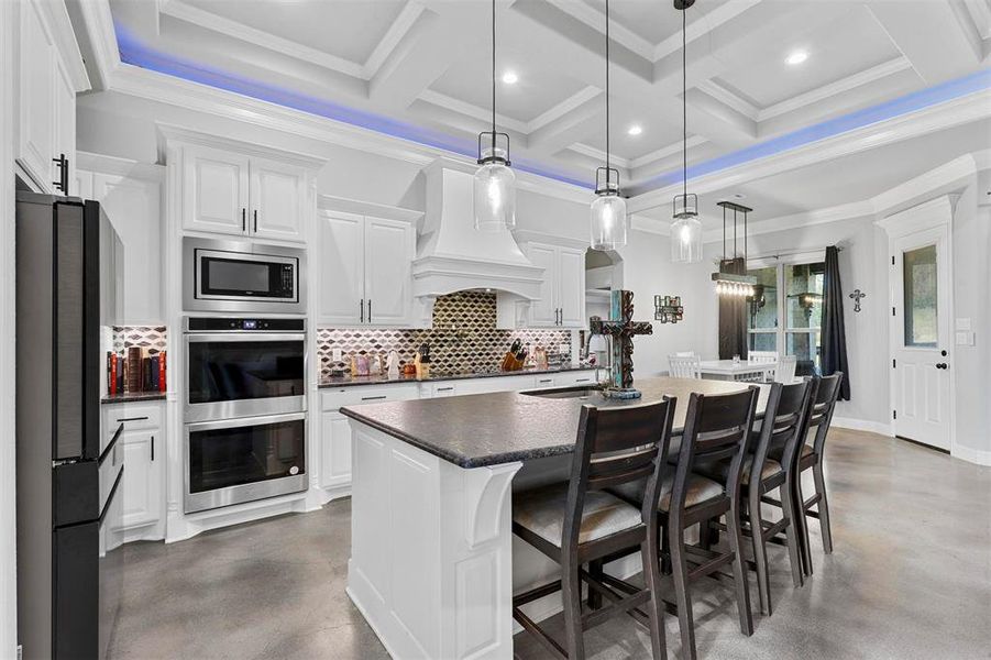 Kitchen with white cabinetry, a kitchen island with sink, stainless steel appliances, decorative light fixtures, and coffered ceiling