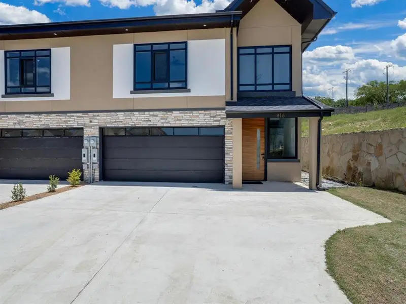 Contemporary exterior featuring a natural wood-finish front door, black-framed windows, stacked stone accents, and a concrete driveway Contemporary exterior featuring a natural wood-finish front door, black-framed windows, stacked stone accents, and a concrete driveway