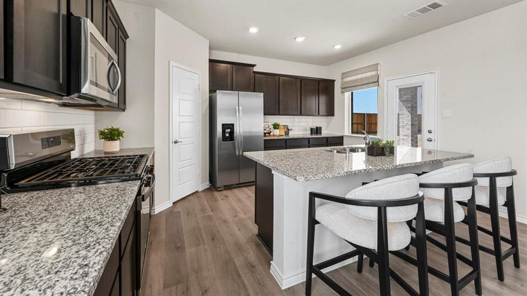 Kitchen featuring appliances with stainless steel finishes, light stone countertops, a center island with sink, a kitchen bar, and dark wood-type flooring Kitchen featuring appliances with stainless steel finishes, light stone countertops, a center island with sink, a kitchen bar, and dark wood-type flooring