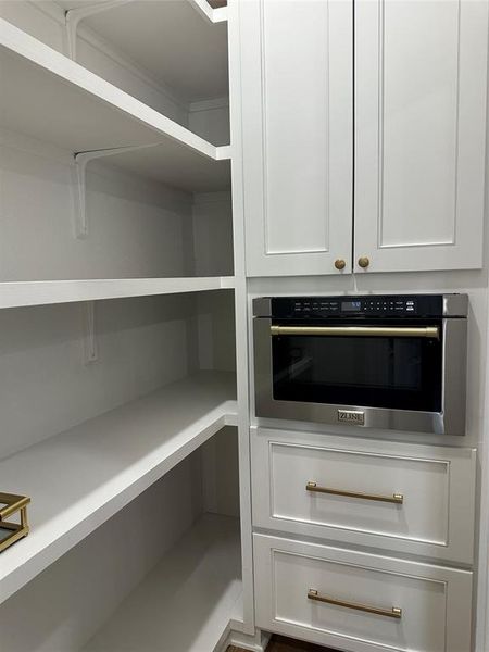 Built-in stainless steel microwave oven, white shaker-style cabinetry with gold-tone hardware, and extensive white shelving