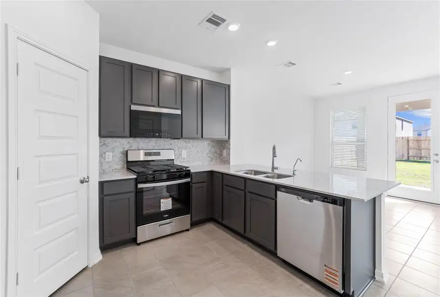 This spacious kitchen, with its high ceilings, showcases Woodmont Dakota cabinets in a Black Fox stain, complemented by Silestone Helix countertops and a modern Daltile backsplash. Stainless steel Whirlpool appliances, recessed lighting, and a pantry complete the space, which offers open sightlines to the expansive Great Room.
