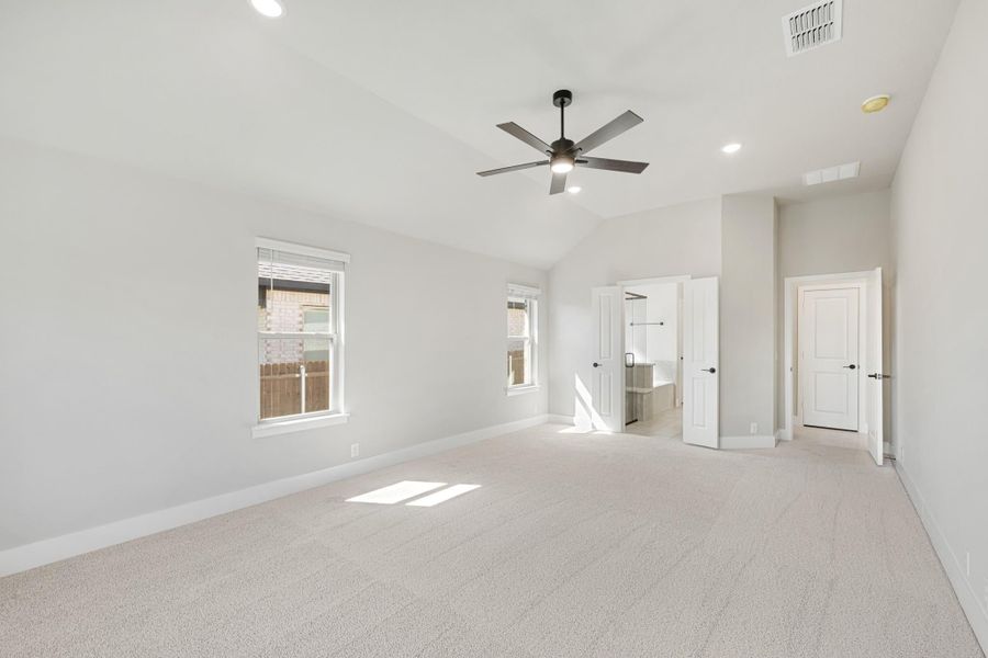 Spacious empty bedroom with vaulted ceiling, ceiling fan, carpet flooring, and doors leading to ensuite bathroom and closet.