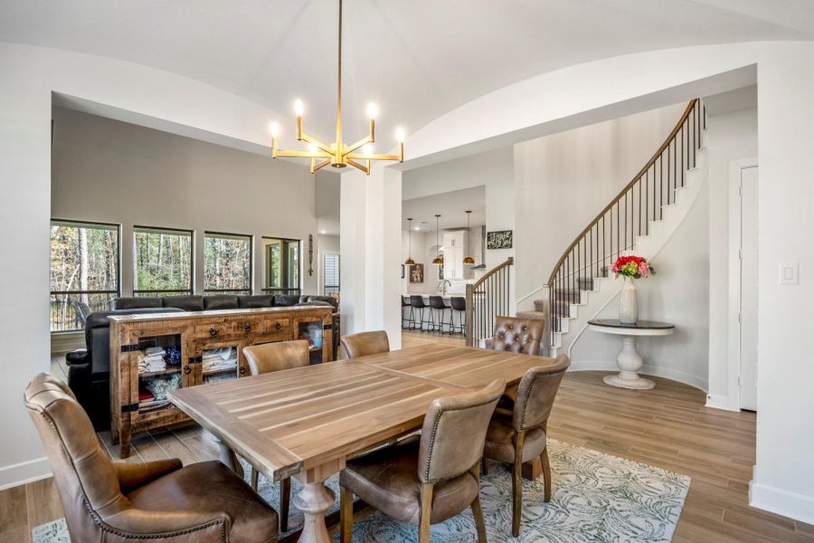 The dining room features a stylish wooden table and chairs under a contemporary chandelier. Large windows provide natural light and views of the outdoors. A curved staircase adds architectural interest, leading to an upper level.